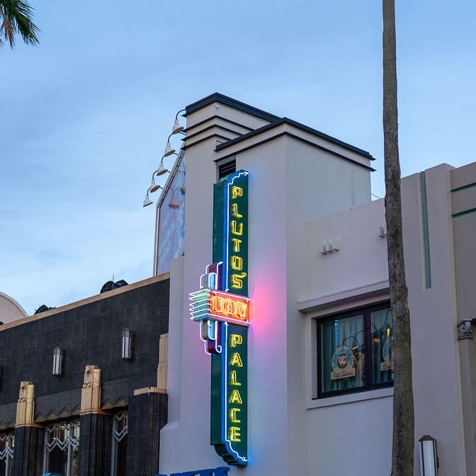A neon sign reading "PLUTO'S TOY PALACE" against a building exterior, with an art deco style and palm trees in the foreground.
