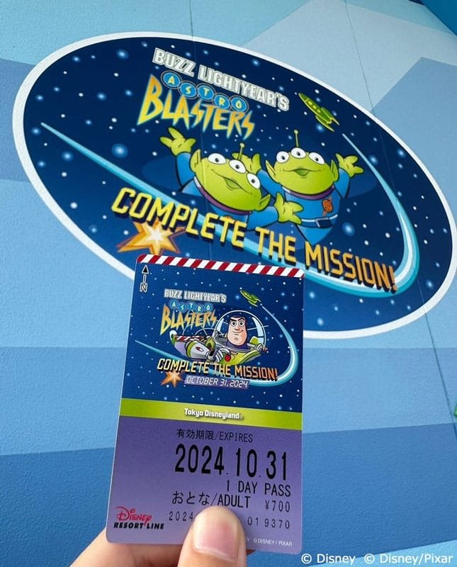 A hand holding a Tokyo Disneyland ticket for "Buzz Lightyear's Astro Blasters," featuring colorful graphics of Buzz Lightyear and green aliens, with the text "COMPLETE THE MISSION! OCTOBER 31, 2024" in a cosmic background. A mural with the same theme is visible in the background.