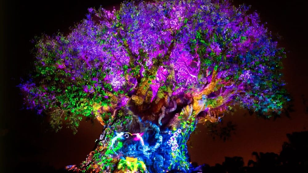 A large, illuminated tree with vibrant purple and green foliage, surrounded by darker shadows, creating a magical, colorful nighttime atmosphere.