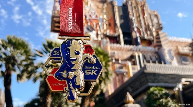 A gold medal featuring a cartoon character resembling Groot from Marvel, with text "2025 Disneyland 5K" and a red ribbon, set against a blurred background of a theme park building and palm trees.