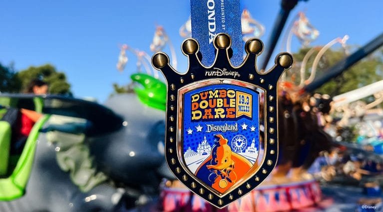 A close-up of a medal hanging from a blue ribbon, featuring the text "DUMBO DOUBLE DARE 19.3 MILES Disneyland" and an illustration of Dumbo, with a blurred amusement ride in the background.