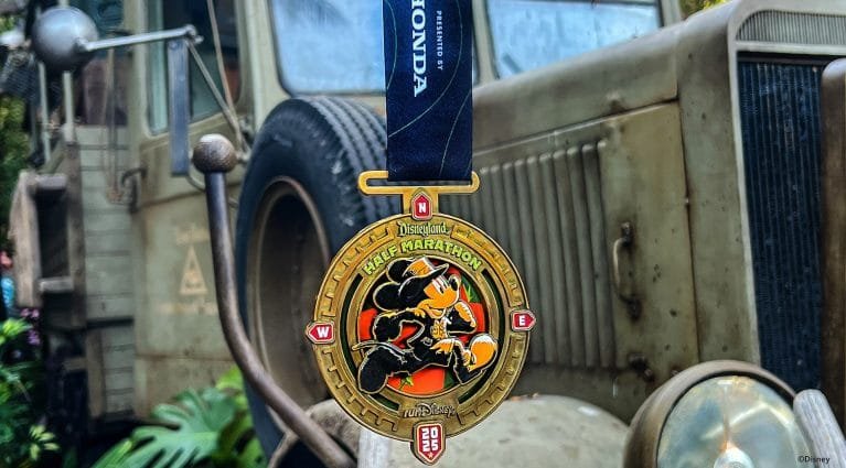 A gold medal hanging with a blue ribbon labeled "Disneyland Half Marathon," featuring an illustration of a cartoon character in running attire. The background shows part of an old vehicle.