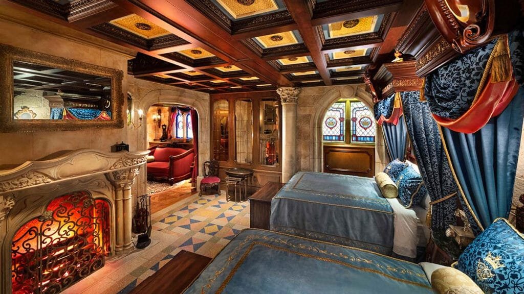 A lavishly decorated bedroom featuring two blue canopied beds, an ornate fireplace with a decorative screen, and stained glass windows. The ceiling has intricate woodwork, and elegant furnishings include a red sitting area and a wooden wardrobe. Rich fabrics and colors dominate the space, giving it a luxurious ambiance.