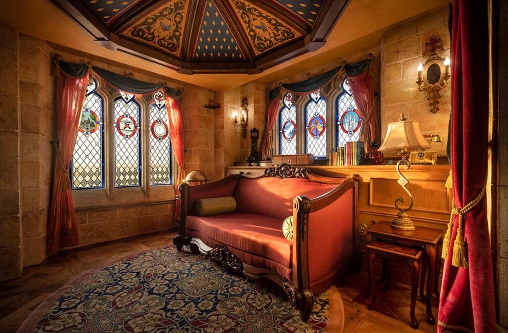 A cozy, elegantly decorated room featuring ornate stained glass windows, a red upholstered sofa, a small wooden table with a lamp, and a richly patterned rug on a wooden floor. The walls are made of stone, and the ceiling has intricate designs.
