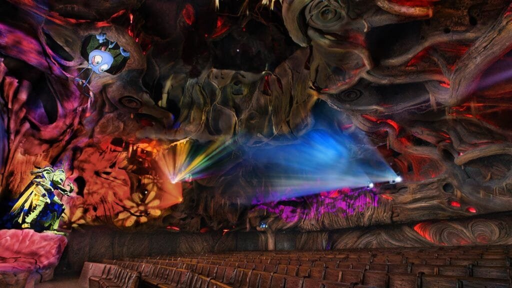 A colorful, cave-like theater interior with intricate, swirling decorations on the walls and ceiling, illuminated by vibrant stage lights. A prominent animated character figure is positioned on a rock formation, while rows of wooden seating face the stage.