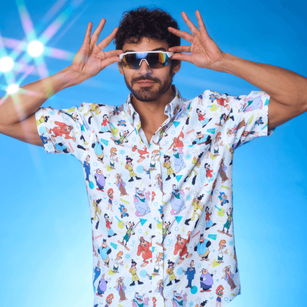A man with curly hair and a beard is striking a playful pose against a blue background, wearing a colorful short-sleeved shirt covered in various animated characters and stylish sunglasses.