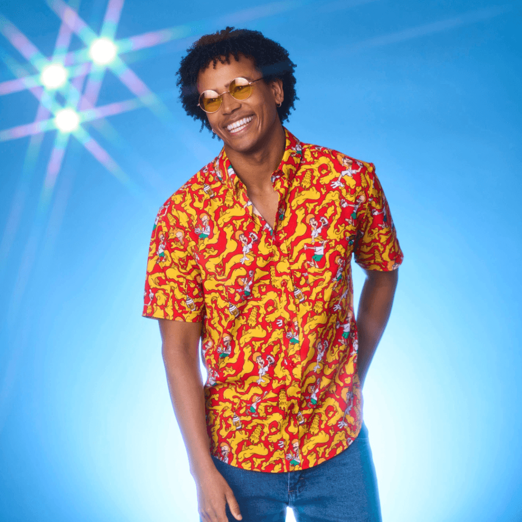 A smiling person with curly hair wearing round yellow sunglasses and a vibrant red shirt adorned with colorful cartoon patterns, standing in front of a blue background with bright light effects.