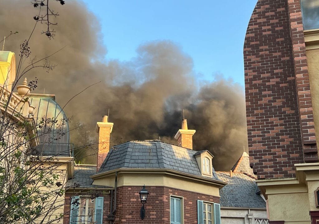 Thick black smoke billows from the rooftops of buildings, partially obscured by a clear blue sky; the foreground features brick and slate-roofed structures with decorative details.