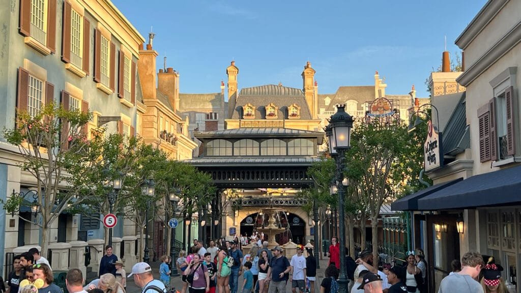 A bustling street view in a themed area, featuring tall buildings with decorative architecture and colorful facades under a clear blue sky. A crowd of visitors fills the walkway, some wearing hats and costumes, while trees and lampposts add greenery and ambiance to the scene.