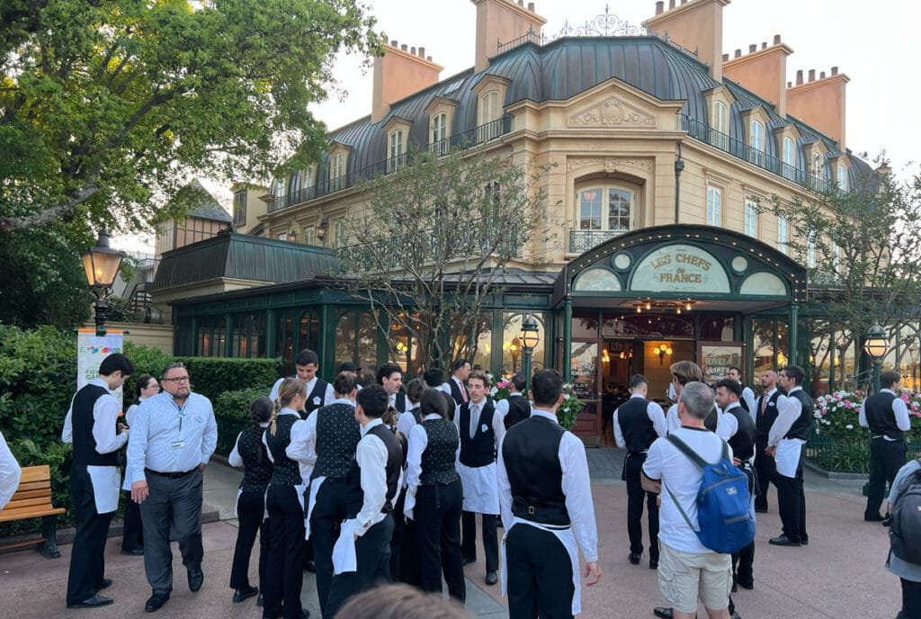A group of waitstaff in formal attire gathers outside the restaurant Les Chefs de France, featuring a building with a classic French design and a green glass entrance surrounded by plants.