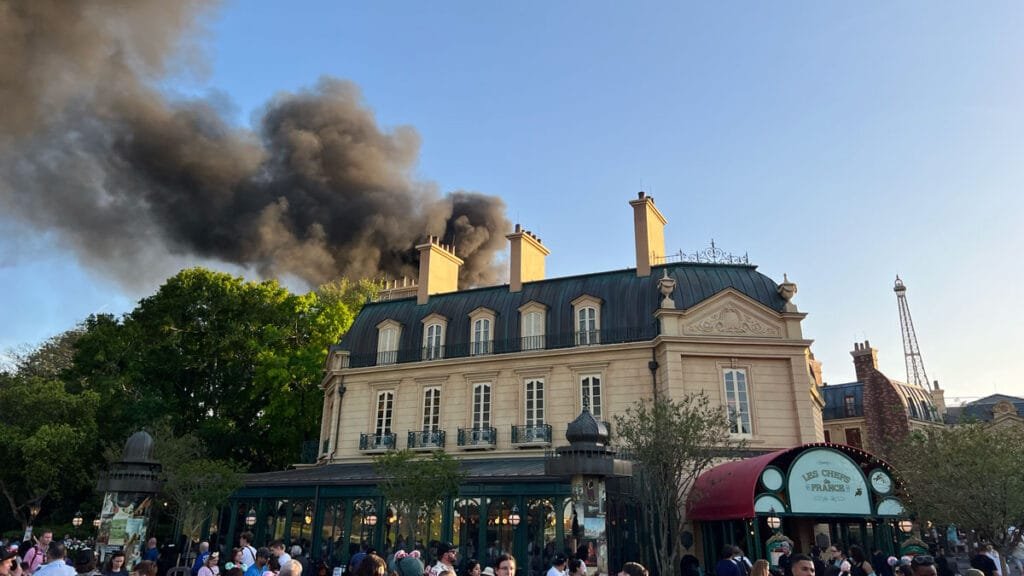 A large building resembling French architecture is shown with a greenish roof and multiple ornate architectural features, while a plume of dark smoke rises above it against a blue sky. Crowds of people gather in the foreground.