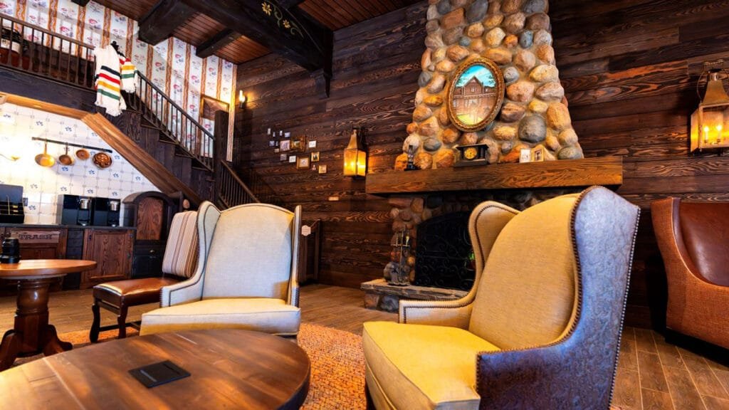 Cozy living room featuring a stone fireplace with a framed painting above it, two upholstered armchairs, a wooden coffee table, and a staircase in the background. The walls are adorned with patterned wallpaper and photographs.
