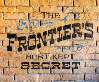A brick wall with a painted sign that reads 'THE FRONTIER'S BEST KEPT SECRET' in bold black letters with decorative blue swirls around it.