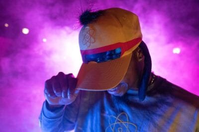 A person wearing a yellow cap with a red stripe and a visor, adorned with a graphic of an atom, is partially obscured by smoke in shades of purple. They hold the cap with one hand, while wearing a gray shirt featuring a similar atom design.