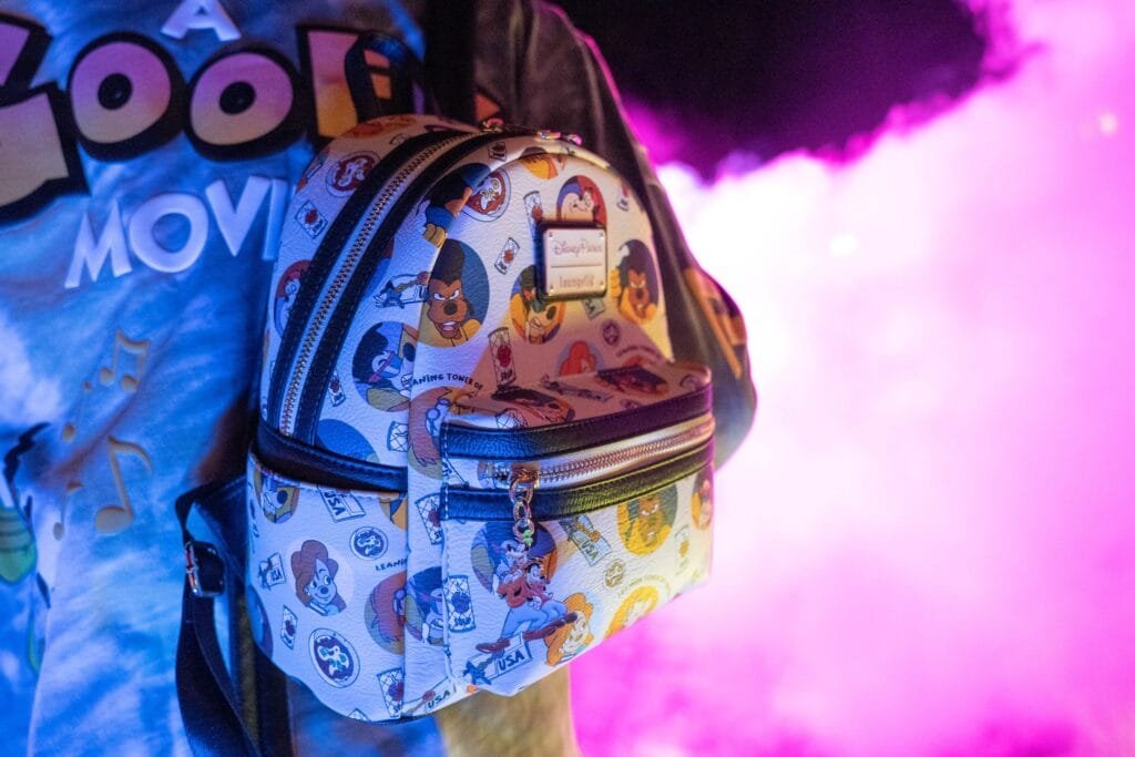 A colorful Disney-themed mini backpack featuring various character patterns, positioned against a backdrop of purple and blue lighting, with part of a shirt that reads 'A Goofy Movie' visible in the background.