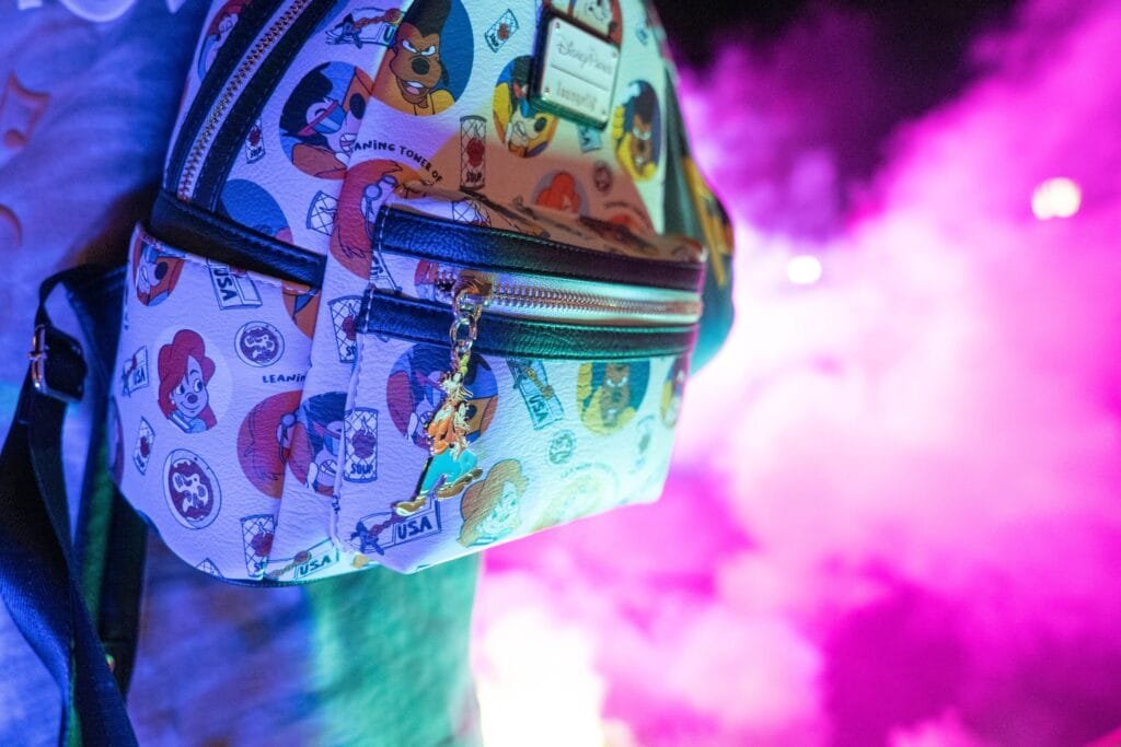 A colorful backpack featuring cartoon character designs, held by a person against a backdrop of vibrant pink smoke and soft lighting, with a keychain accessory visible on the zipper.