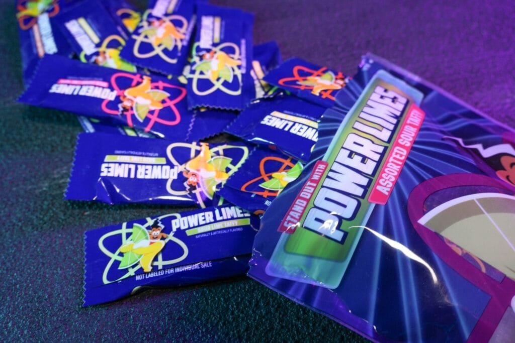 A group of colorful blue wrappers for Power Lime sour taffy scattered on a surface, with a larger pouch of assorted sour taffy in the background, featuring bright graphics and cartoonish characters.