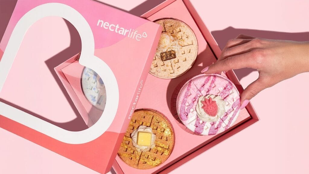 A hand with pink nails reaches toward a pink box of three decorative, colorful desserts resembling waffles, with one topped with a small strawberry and others adorned with icing and glitter, against a light pink background.