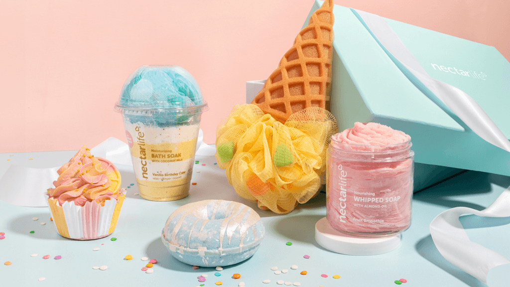 An array of colorful bath and body products displayed on a light blue surface, including a cupcake-shaped bath bomb, a moisturizing bath soak in a transparent cup, a yellow loofah, a blue and white donut-shaped bath bomb, and a jar of whipped soap, all set against a pastel pink background and a light teal box.