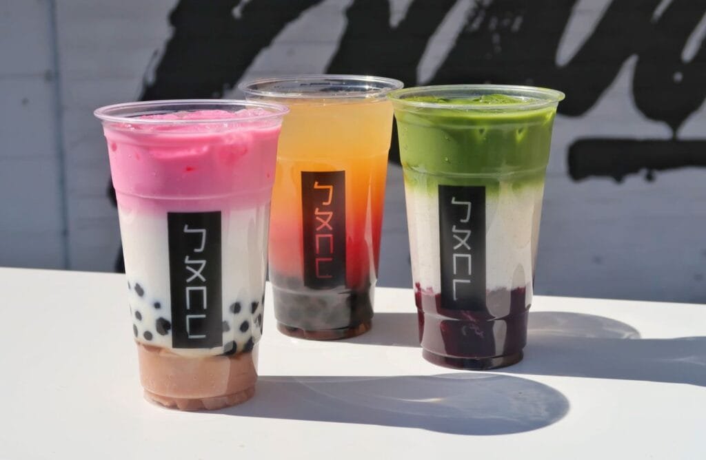 Three colorful drinks in clear cups on a table, with a pink and white drink on the left featuring black tapioca pearls, a yellow drink in the center, and a green layered drink on the right, all displaying a black label with a logo or text in the front.