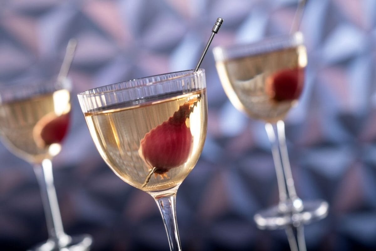 Three cocktail glasses filled with a clear liquid and garnished with pickled onions, set against a blurred geometric background.