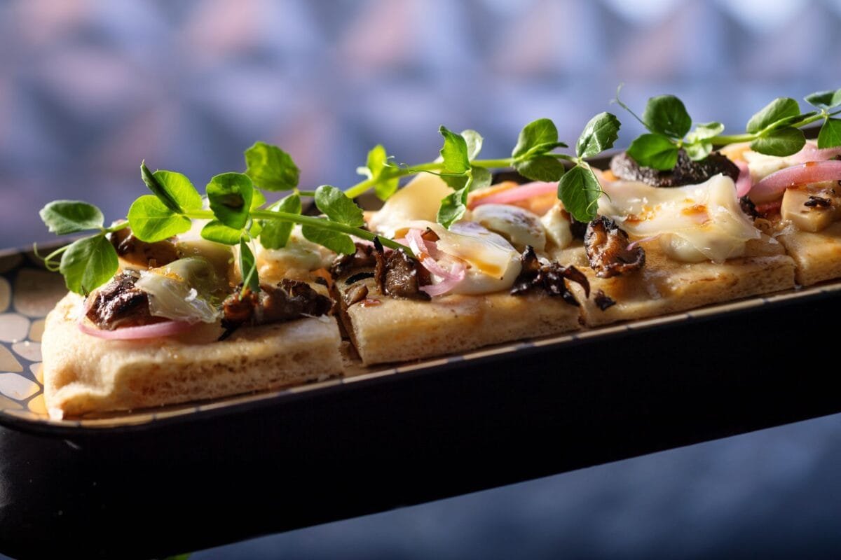 Sliced gourmet flatbread topped with melted cheese, mushrooms, onions, and fresh green garnishes, served on a decorative platter.