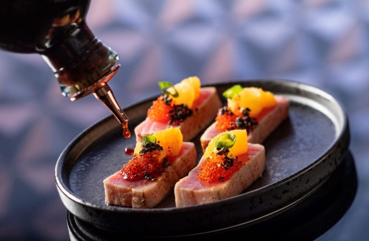Slices of sushi-grade tuna topped with roe, orange segments, and garnished with green leaves, being drizzled with soy sauce from a bottle, served on a black plate.