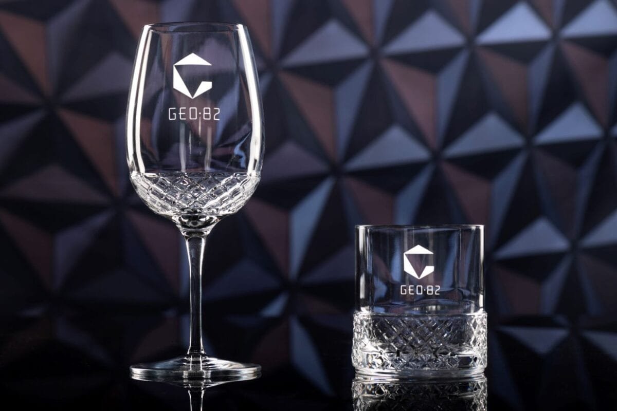 A clear, intricately cut wine glass and a short tumbler glass, both featuring the logo "GEO:82," against a geometric-patterned background.