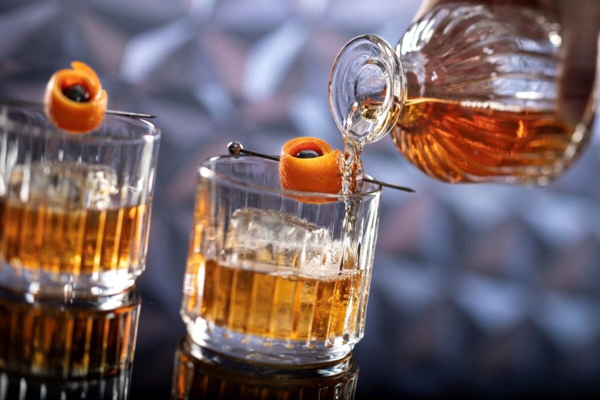 A close-up view of two whiskey glasses with ice cubes, one being filled with whiskey from a decanter. Each glass is garnished with an orange peel and a dark cherry on a metal skewer.