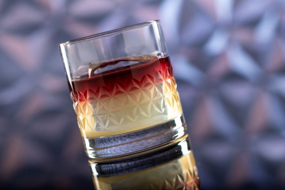A close-up of a layered cocktail with a deep red top and a light yellow bottom in a geometric-patterned glass, set against a blurred background.