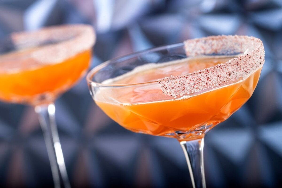 Close-up of two orange cocktails in decorative glasses with sugared rims, set against a blurred geometric background.