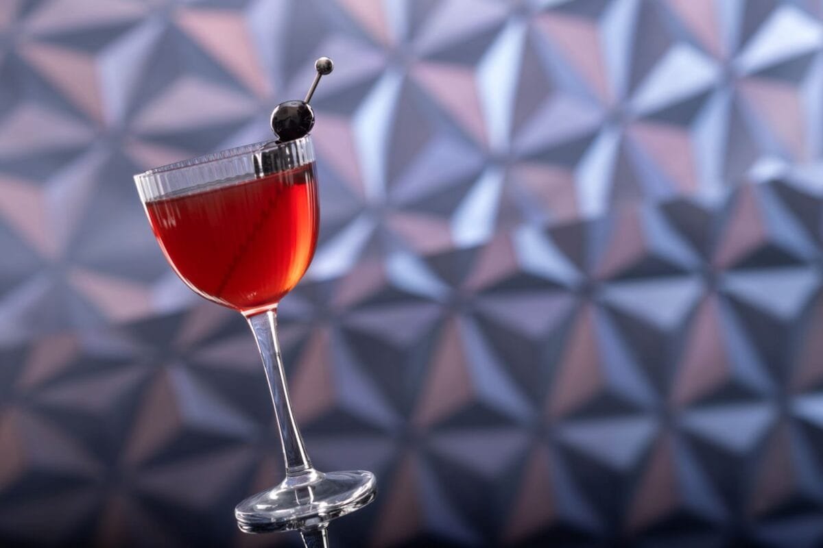 A red cocktail in a stemmed glass garnished with a cherry, with a geometric patterned background.