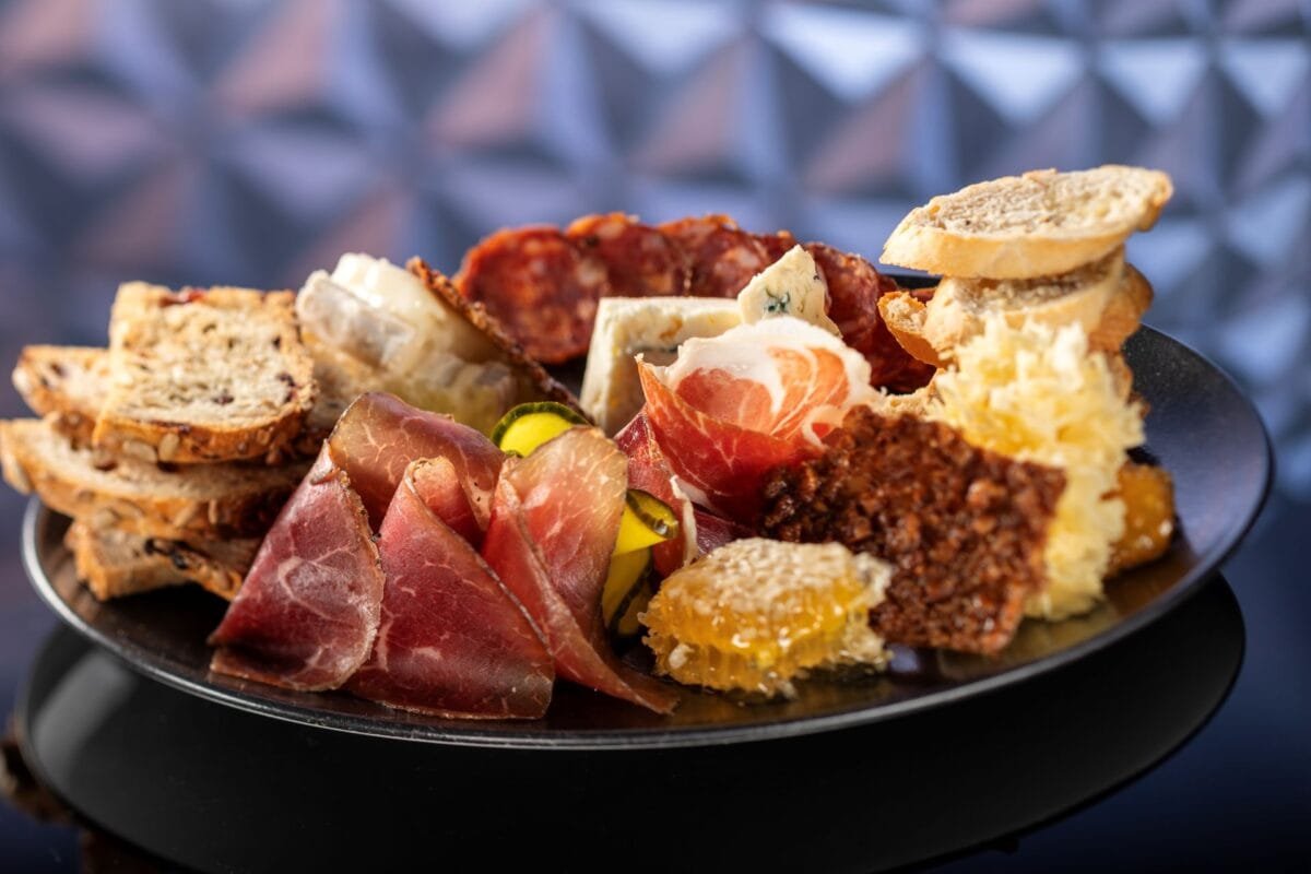A charcuterie board featuring assorted cured meats, slices of bread, cheese, pickles, and honeycomb, elegantly arranged on a black plate.