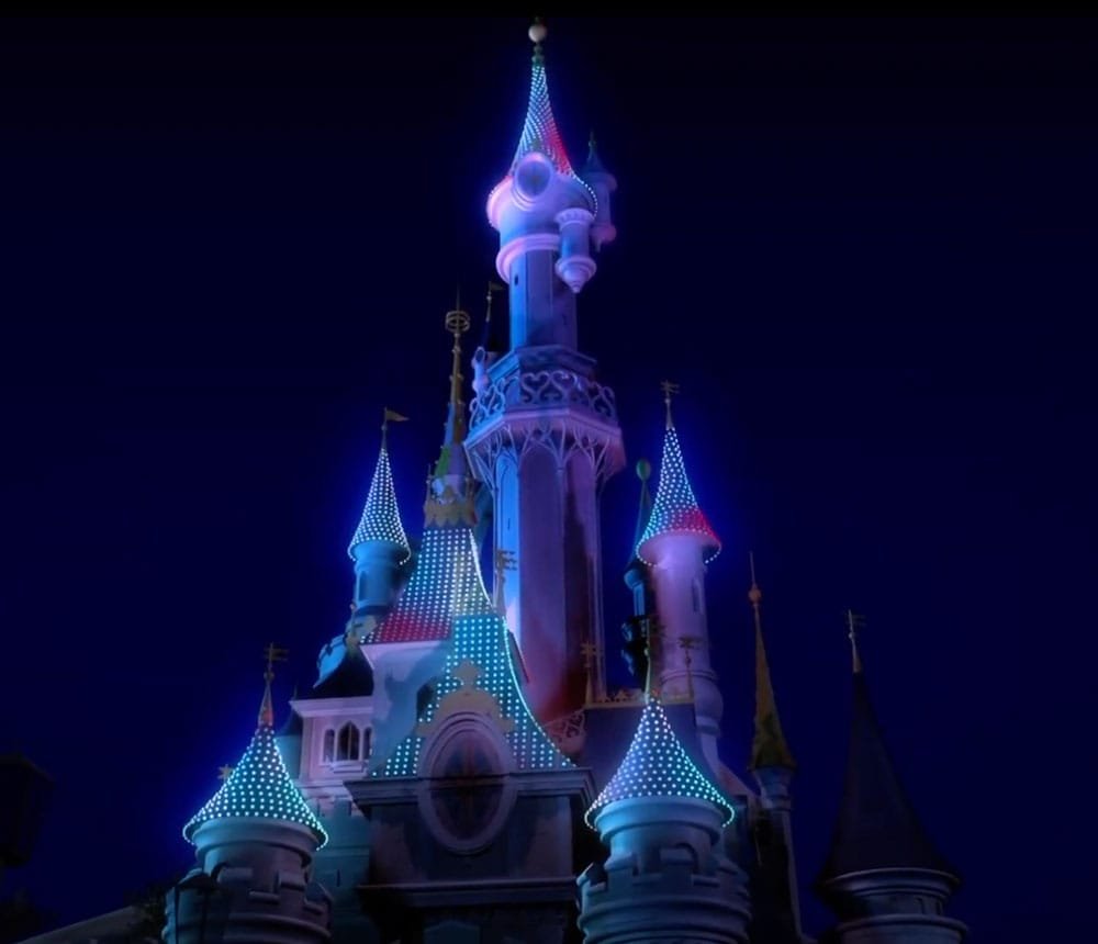 A illuminated castle at night, featuring multiple turrets with colorful lights in blue, pink, and white, set against a dark sky.