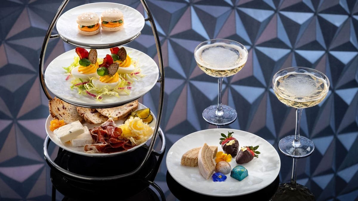 A three-tiered serving tray featuring an assortment of gourmet foods including macarons, deviled eggs with garnish, cheese, cured meats, and bread, alongside a plate of desserts such as chocolate-covered strawberries, chocolates, and pastries. Two champagne glasses are filled with sparkling wine, set against a geometric patterned background.