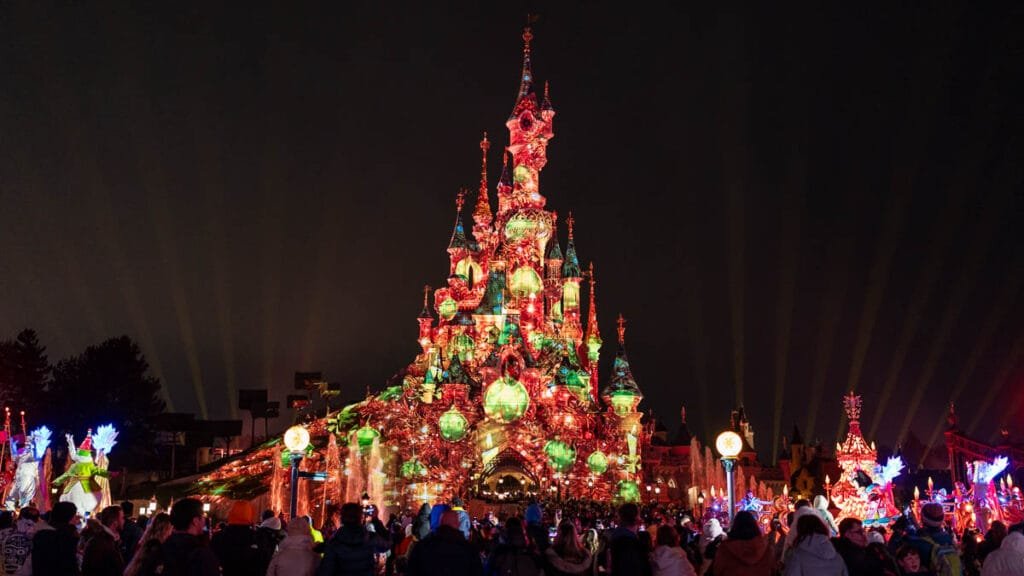 A brightly lit fairytale castle adorned with colorful lights and decorations at night, surrounded by a crowd of people admiring the display, with illuminated fountains and whimsical figures in the foreground.