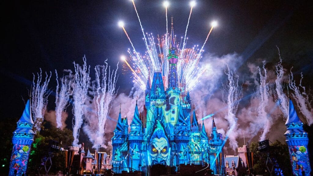 A vibrant castle illuminated in blue light with a large figure projected on its facade, surrounded by colorful fireworks and smoke against a night sky.