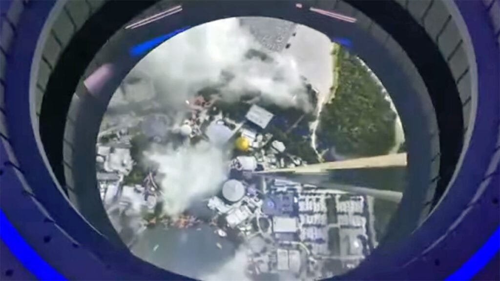 Aerial view of a coastal city with buildings and green spaces, partially obscured by clouds, as seen through a circular window from a spacecraft or a high-altitude viewing platform.