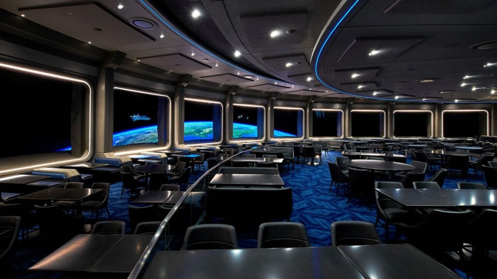 A futuristic dining room with circular seating and tables, featuring large windows displaying a view of Earth from space with a dark background. The room has blue ambient lighting and a sleek, modern design.