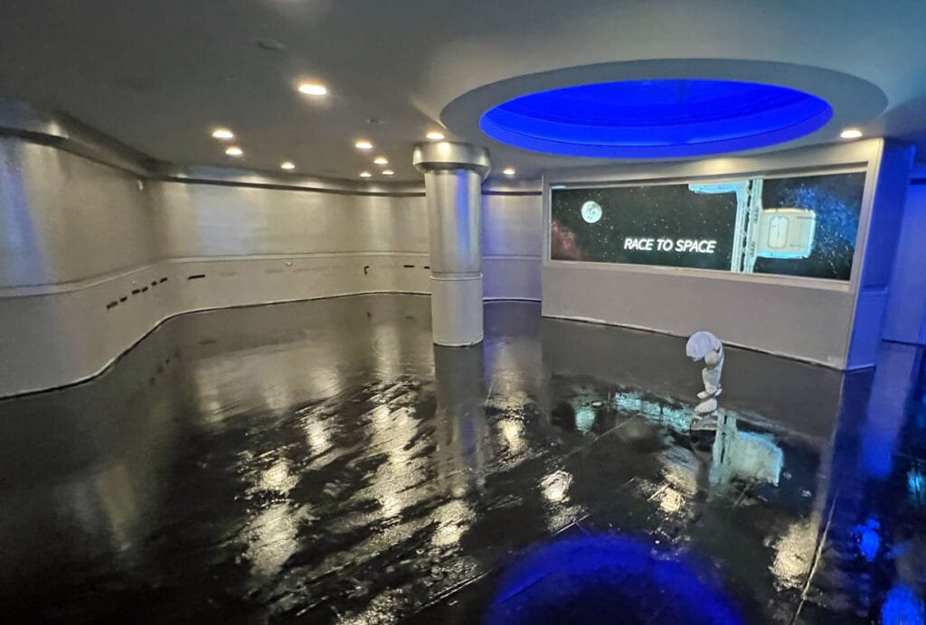 An interior space featuring shiny silver walls and a glossy black floor, with blue lighting above. A large screen displays the text 'RACE TO SPACE,' and a small astronaut figure is positioned on the floor.